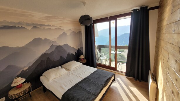 A bedroom with a mountain mural, a bed, a small table with a lamp, and a view of the outdoors through a large window.