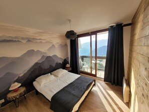 Une chambre avec une fresque représentant une montagne, un lit, une petite table avec une lampe et une vue sur l’extérieur grâce à une grande fenêtre.