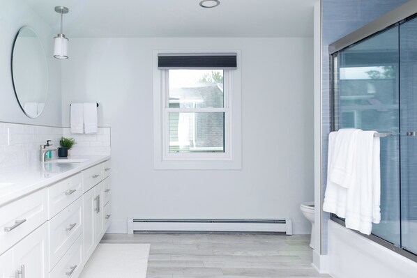 A modern bathroom with a white vanity, a large mirror, a window, and a glass shower enclosure.