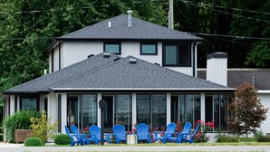 A two-story house with a large front porch, a covered outdoor seating area with blue chairs, and a stone retaining wall in front of a body of water. - Modern Lakefront Gun Lake Cottage Near Bay Pointe Inn (Shelbyville)