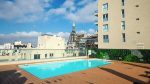 Uma piscina no terraço com deck de madeira, vista de um edifício histórico com pináculo verde e um prédio de apartamentos moderno ao fundo.