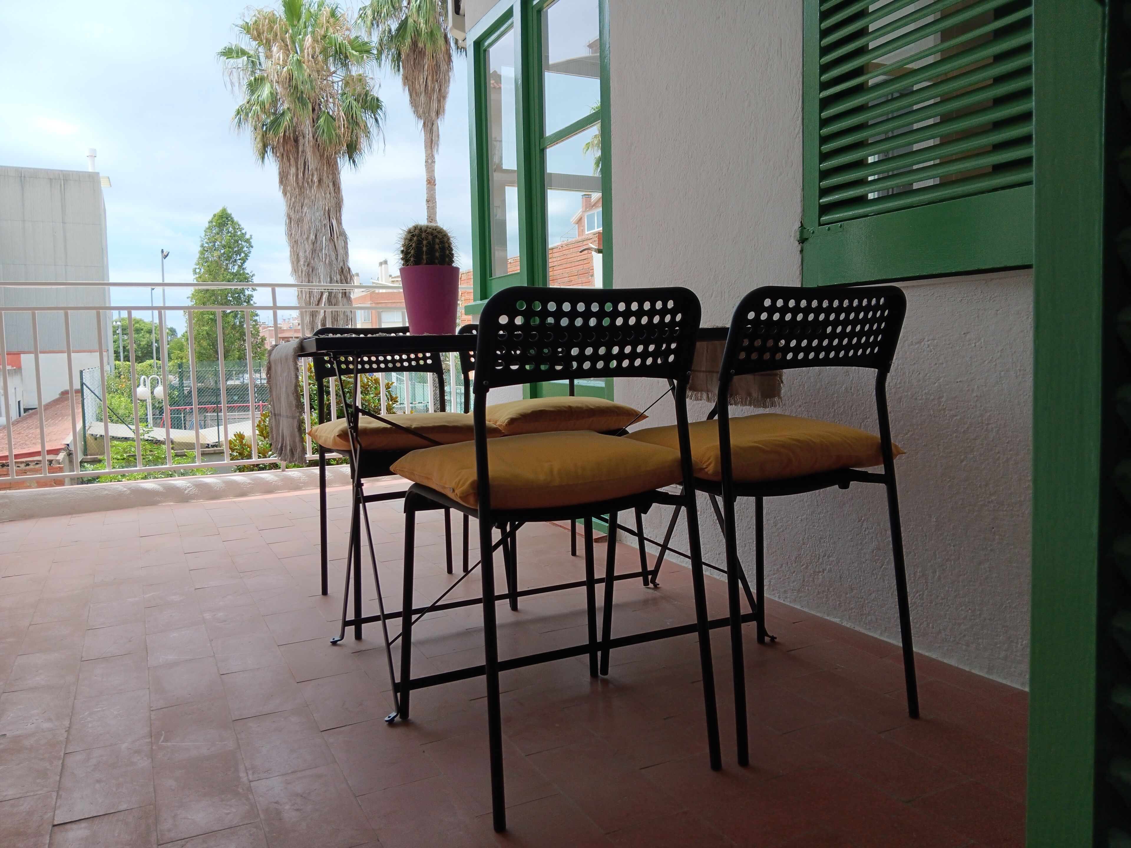 A balcony with two black metal chairs with yellow cushions, a potted plant, and a view of a building and palm trees.