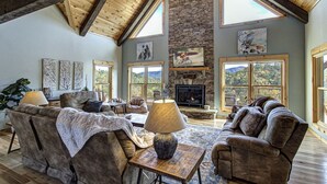 A spacious living room with a stone fireplace, large windows, and a high wooden ceiling. - Luxury Smoky Mountain Cabin | Amazing Views, Hot Tub, Fire Pit Near Pigeon Forge (Sevierville)