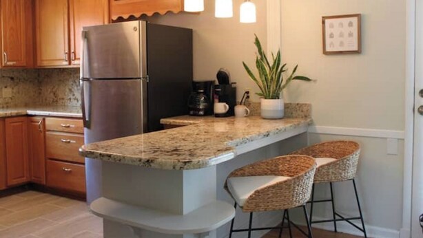 Una cocina con armarios de madera, un refrigerador de acero inoxidable, encimera de granito, dos taburetes de bar, una planta en maceta e iluminación colgante.