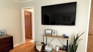 Una sala de estar con una televisión de pantalla plana montada en la pared, un aparador de madera, una cama para mascotas, una mesita con un marco de fotos y una vela, y una planta en maceta.