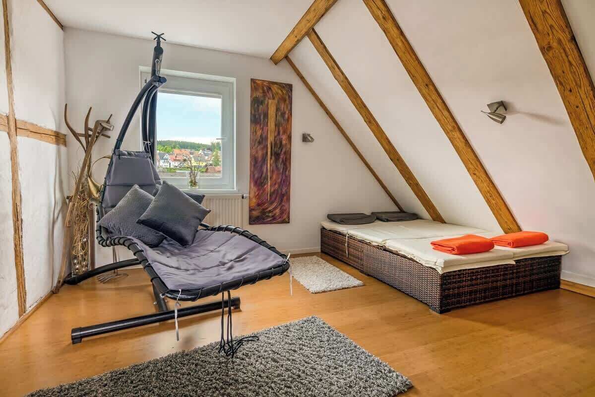 Un dormitorio tipo loft con una hamaca colgante, una cama con almohadas naranjas, un ventanal con vistas y un cuadro abstracto colorido.