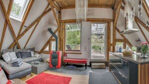 Ein Loft-Zimmer mit Holzbalkendecke, einer Couch, einer Bank, einer Küchenzeile und Blick durch ein großes Fenster.