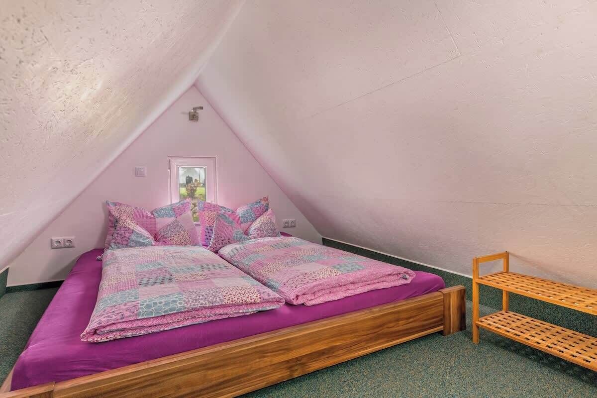 A small, cozy attic bedroom with a wooden bed, a wooden bench, and a window with a view of greenery.