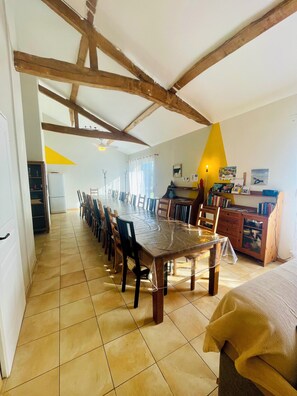 A spacious dining area with a long wooden table, chairs, and a kitchen area with cabinets and a refrigerator.
