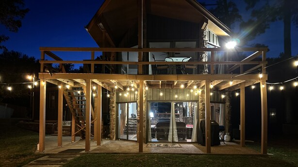 A two-story wooden house with a covered patio area, string lights, and a staircase.