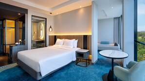 Quarto de hotel moderno com uma cama grande, uma mesinha de cabeceira, uma mesa redonda pequena e vista para o exterior.