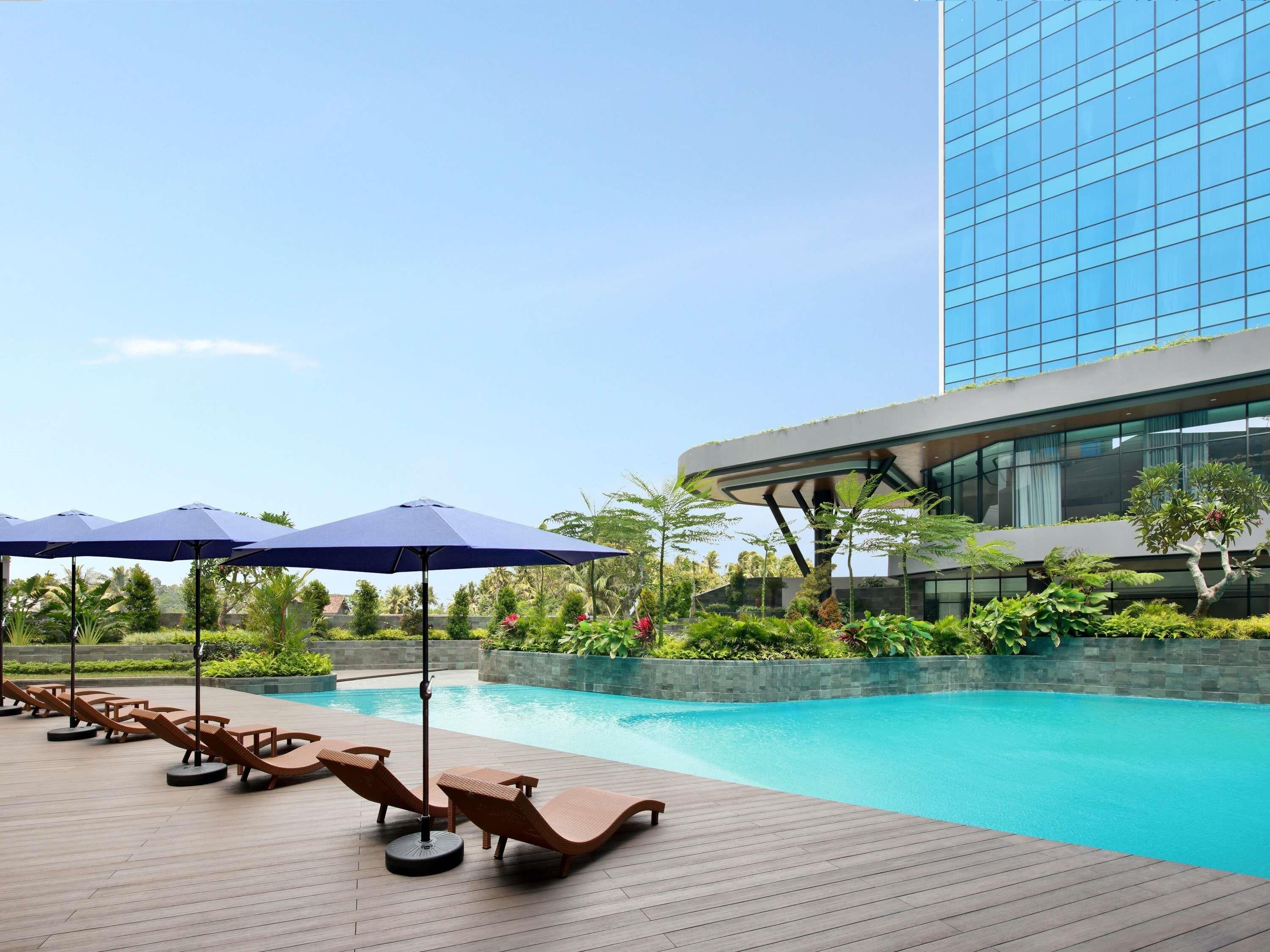 Un área junto a la piscina con sillones de descanso y sombrillas, rodeada de vegetación y un edificio moderno.