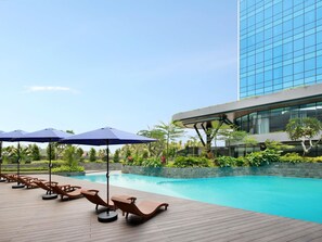 A poolside area with lounge chairs and umbrellas, surrounded by greenery and a modern building. - Novotel Yogyakarta International Airport Kulon Progo (Temon)
