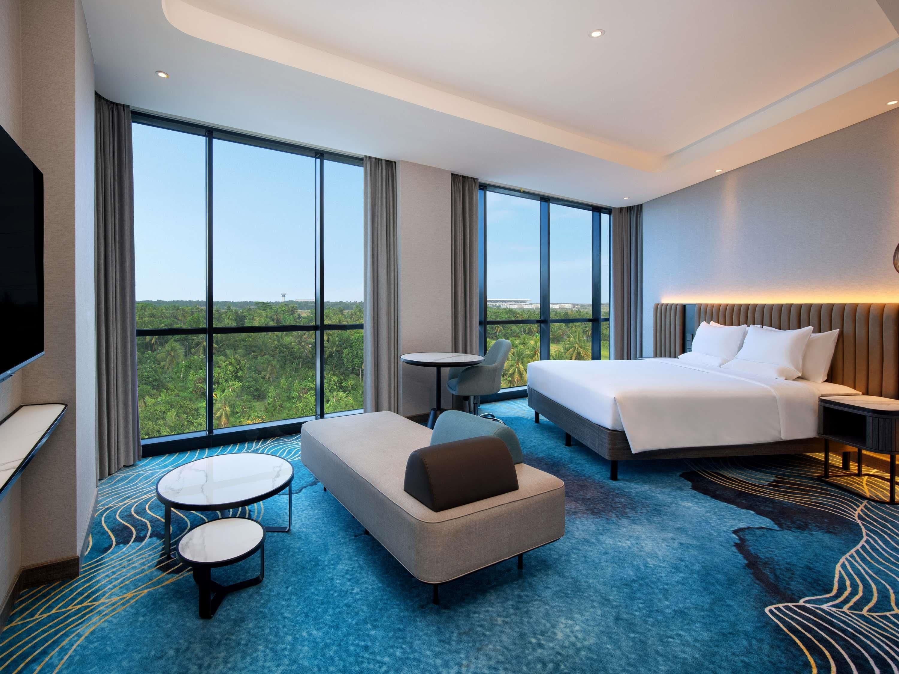 Habitación de hotel con una cama grande, un sofá, una pequeña mesa, un televisor y vistas a zonas verdes a través de amplios ventanales.