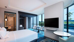 Quarto de hotel moderno com uma cama grande, televisão, área de estar com mesa e cadeiras, e vista para o exterior.