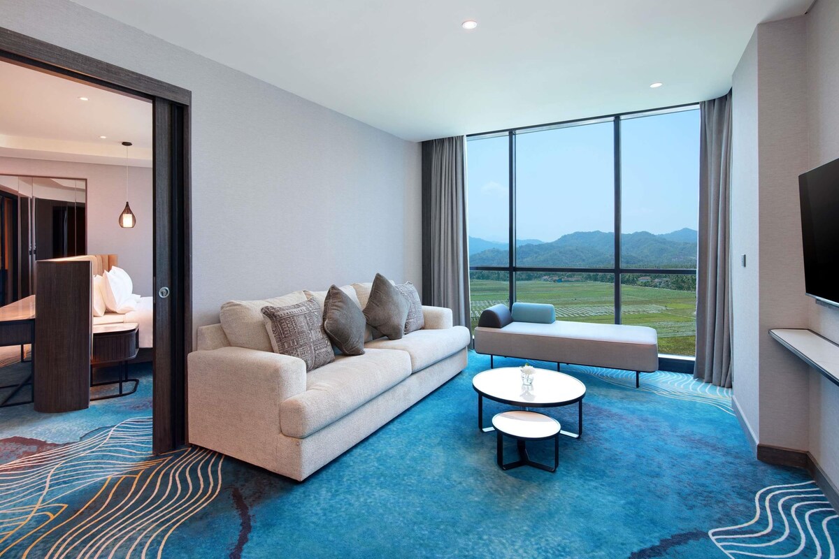 A modern hotel room with a sofa, a small table, and a view of a mountainous landscape through large windows.