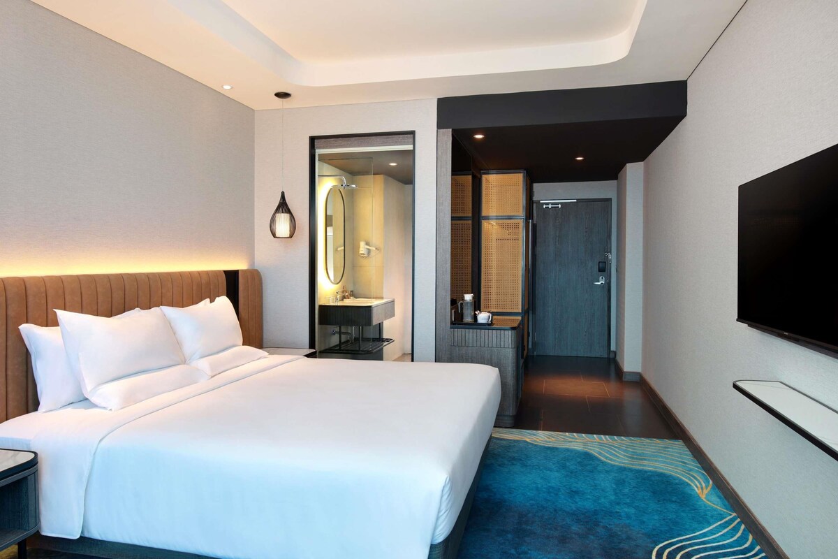 A modern hotel room with a large bed, a bathroom with a mirror and sink, and a flat-screen TV mounted on the wall.