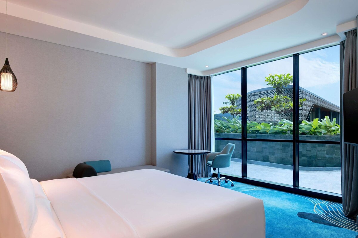 A hotel room with a bed, a small table, a chair, and a large window offering a view of the outdoors.