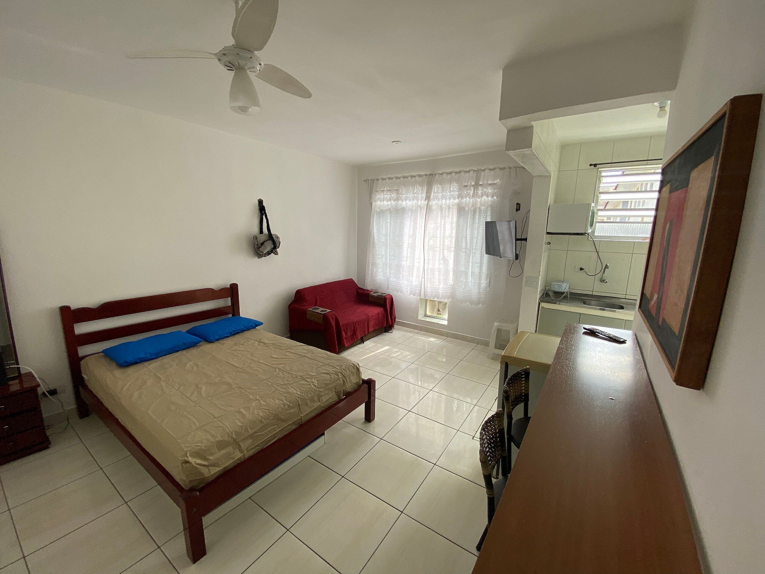 Une chambre avec un lit, une chaise rouge, un ventilateur de plafond, une fenêtre avec des rideaux et un coin cuisine visible à travers une porte ouverte.