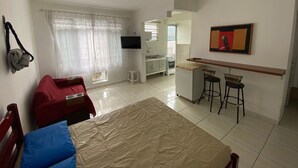 A compact hotel room with a bed, a desk, a chair, a TV, and a kitchenette. - Foot in the sand Beach corner of the fort (Praia Grande)
