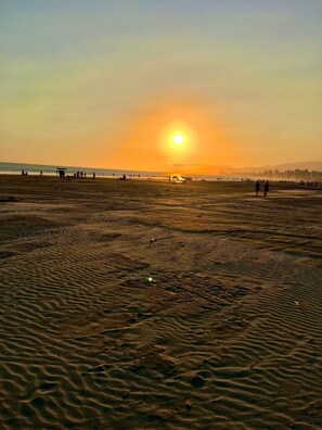 Ein Strand mit untergehendem Sonnenuntergang im Hintergrund, sandigem Untergrund und einigen Personen in der Ferne.