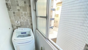 A small balcony with a washing machine, a potted plant, and a view of a modern building. - Foot in the sand Corner of Fort II - Tiradentes 3 (Praia Grande)