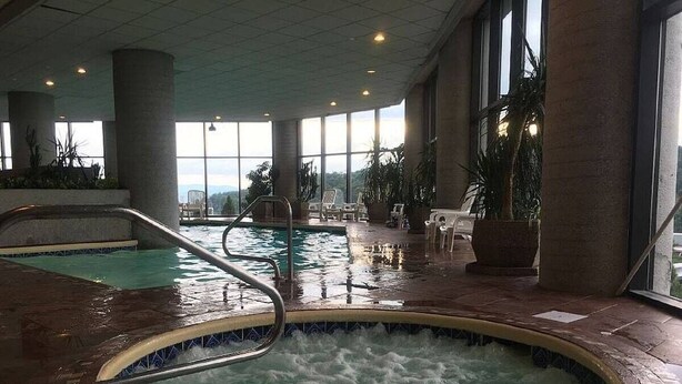 En indendørs swimmingpool med et spabad, omgivet af liggestole og store vinduer.
