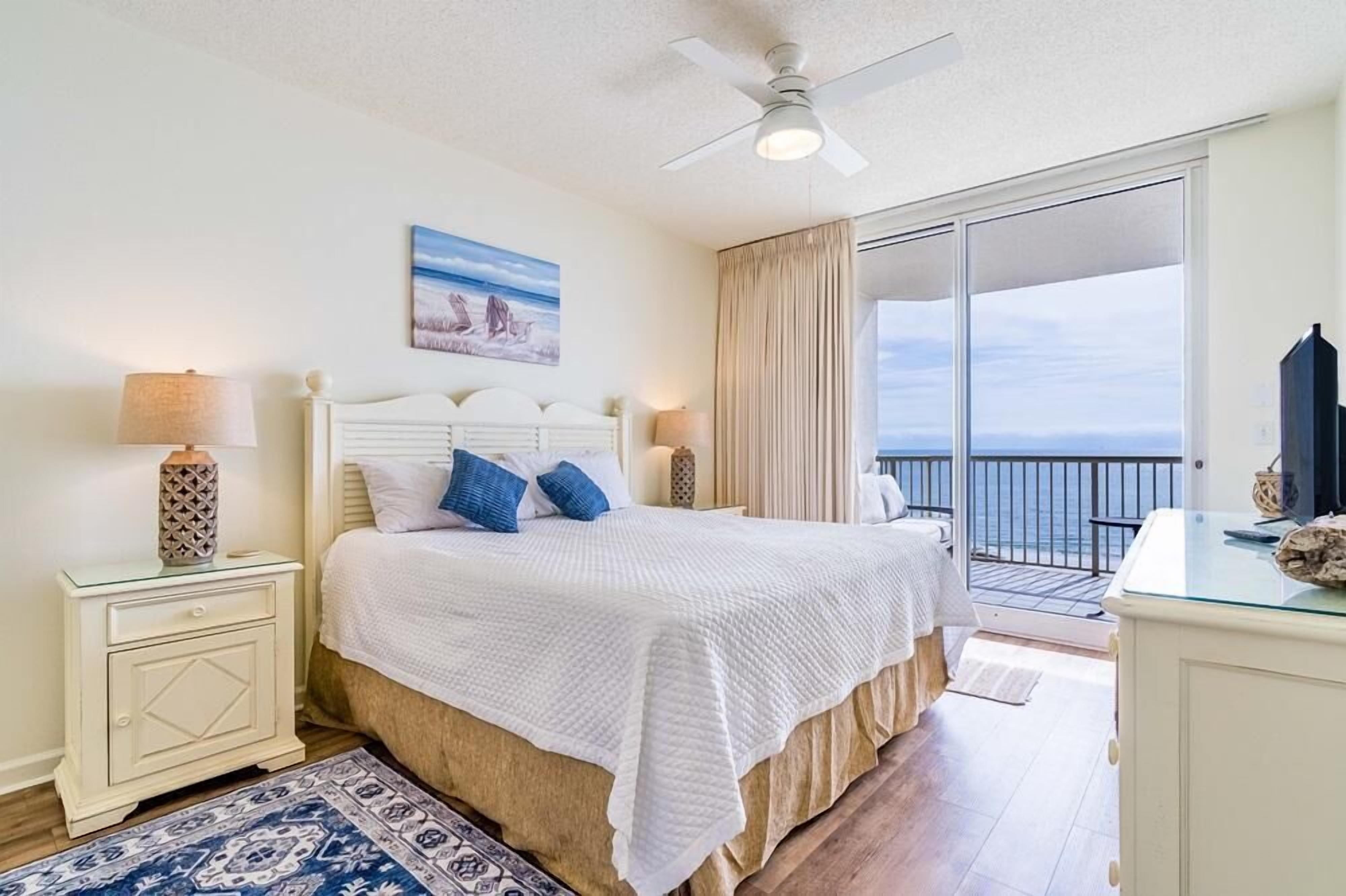 Une chambre à coucher comprenant un lit, des tables de chevet, un ventilateur de plafond, un tableau et une vue sur l’océan, visible à travers une porte coulissante.