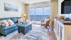 A living room with a sofa, ottoman, and chair, a flat-screen TV, and a view of the ocean through large sliding doors.