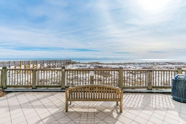 A wooden bench on a tiled deck overlooking a beach with a wooden fence and a trash can.