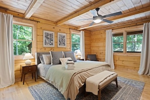 A cozy bedroom with a bed, bedside tables, a ceiling fan, and large windows with curtains.
