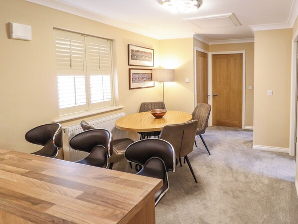 A dining area with a round table, six chairs, a wooden table, a radiator, a window with blinds, a framed picture, a lamp, and a door.