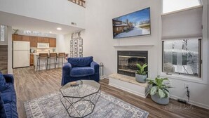 A spacious living room with a fireplace, a kitchen in the background, and a large window.