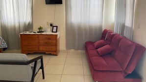 Un salon avec une commode en bois, un canapé rouge, un téléviseur fixé au mur et deux fenêtres ornées de rideaux légers.