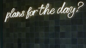 Une enseigne au néon avec l’inscription « quels sont vos projets pour la journée ? » sur un fond sombre à motifs de carreaux.