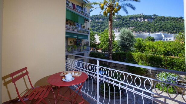 Un balcón con una mesa y una silla rojas, con vistas a un paisaje verde exuberante con una palmera y un edificio a lo lejos.