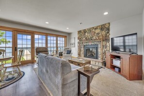 A spacious living room with a stone fireplace, a television, and a view of the outdoors through large windows. - Anchors Away Cottage on Big Spirit Lake (Lakefront) (Orleans)