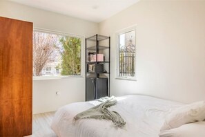 A bedroom with a bed, a window, a metal shelf unit, and a view of trees and a building outside.