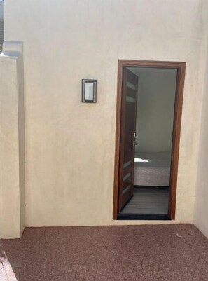 A room with a door leading to a bed, a small window, and a wall-mounted light fixture.