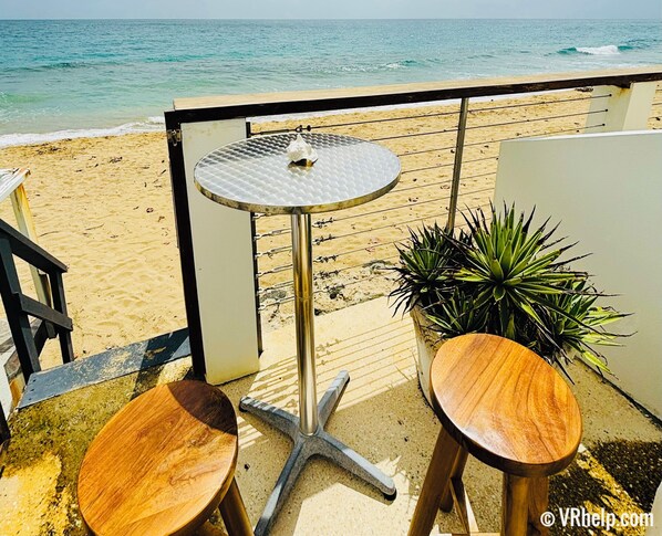 Un balcón con una mesa redonda y dos taburetes, con vista a una playa de arena y al océano.