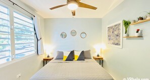 Un dormitorio con cama, dos mesitas de noche, ventilador de techo, tres espejos en la pared, ventana con persianas y repisa con plantas.