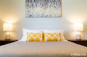 A neatly made bed with white and yellow patterned pillows, two bedside lamps, and a textured abstract painting on the wall.