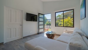 A modern living room with a sofa, a coffee table, a TV, and a large window with a view of greenery. - Cozy house, pool, WIFI, mins to beach, Uvita (Ballena)