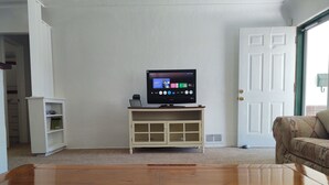 En stue med et træsofabord, et fjernsyn på et hvidt stativ, en beige sofa og en hvid dør.