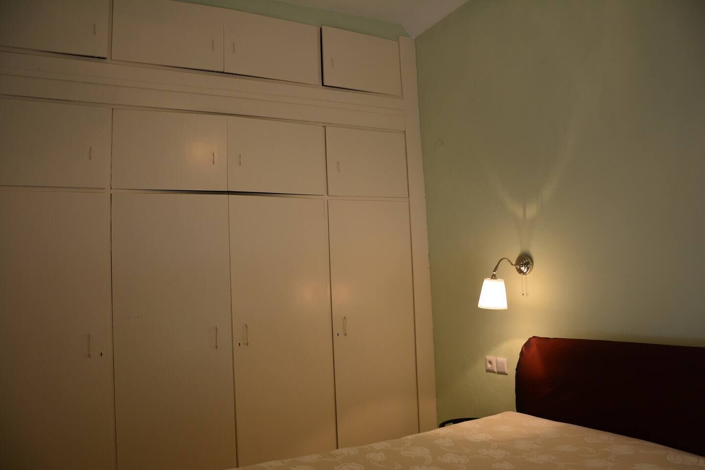 A bedroom with a bed, a nightstand, a wall-mounted lamp, and a wardrobe with multiple doors.