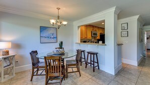A dining area with a round glass table, wooden chairs, a chandelier, and a view into the kitchen. - Palmetto Dunes Golf Getaway: Pool + Beach Access! (Hilton Head Island)