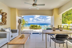 A balcony with a view of the ocean, a ceiling fan, a wooden table, and a sofa.