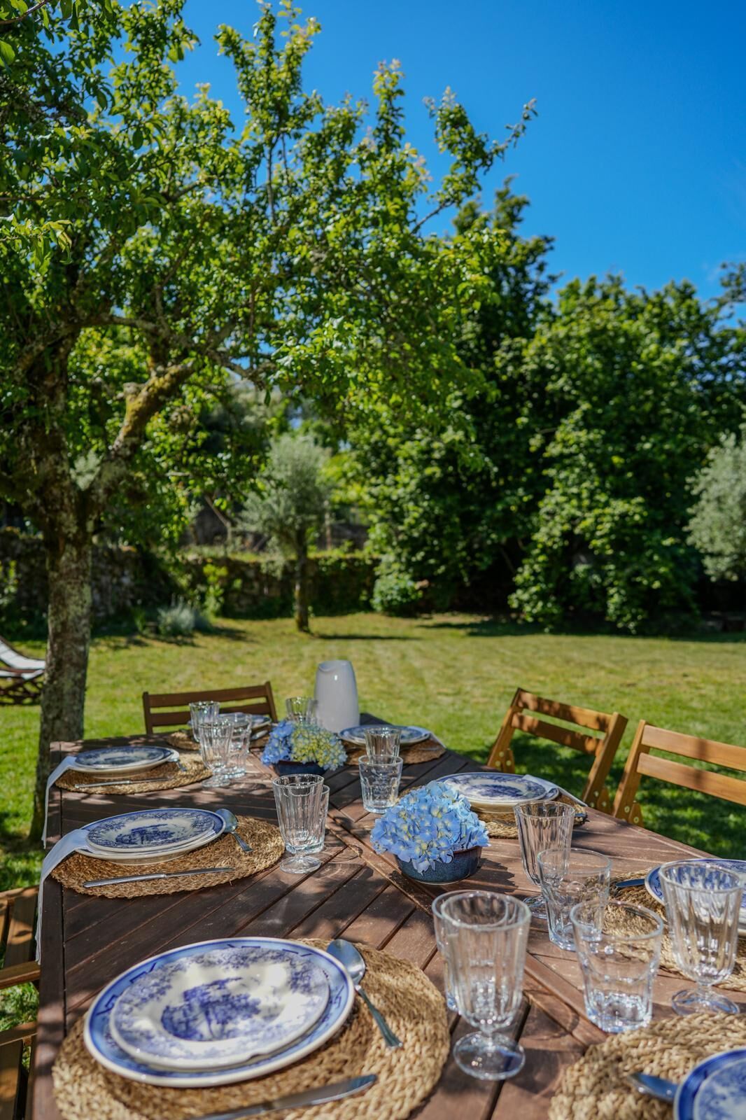 Een buitentafel gedekt met borden, glazen en een karaf, omringd door bomen en een gazon.