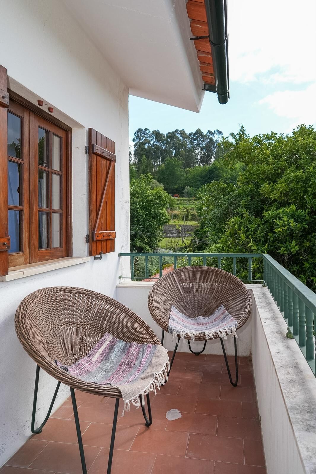 A balcony with two wicker chairs, a window with shutters, and a view of a green landscape.