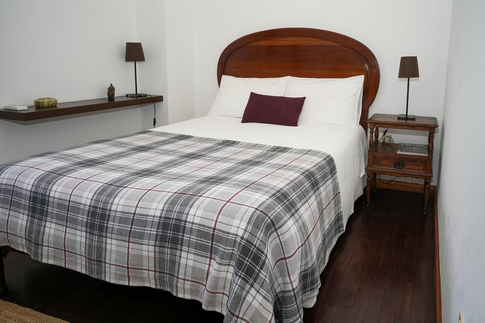 A bedroom with a wooden headboard, a plaid bedspread, two bedside tables with lamps, and a small shelf with decorative items.